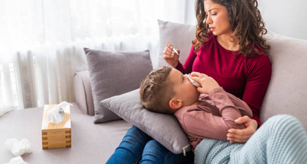 Mother caring for sick child lying on sofa with tissues and thermometer.