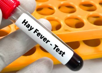 Gloved hand holding a blood sample tube labelled “Hay Fever – Test” beside a test rack