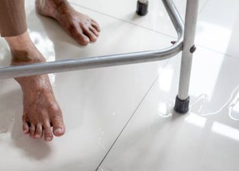 Elderly person with foot drop using a walker beside a puddle on the floor
