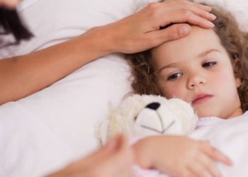 Sick child in bed with a concerned parent checking her forehead for fever