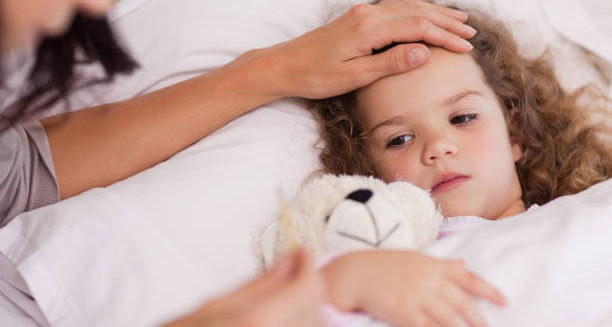 Sick child in bed with a concerned parent checking her forehead for fever