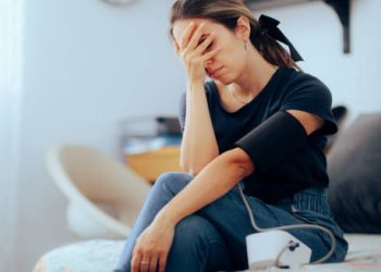 Woman sitting with a blood pressure cuff, appearing dizzy or unwell, indicating low blood pressure.