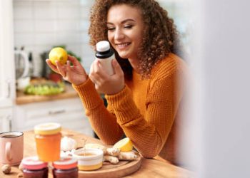 Smiling woman holding supplements and lemon, surrounded by natural remedies in kitchen