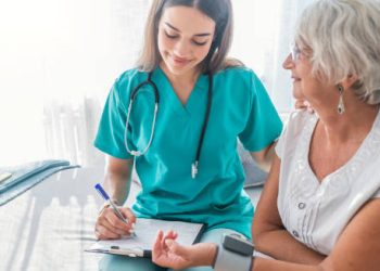 Nurse monitoring elderly woman’s blood pressure and recording results during a home visit