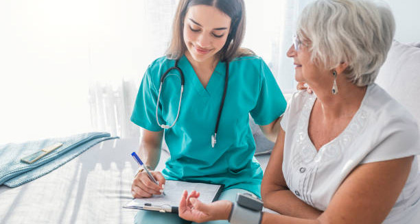 Nurse monitoring elderly woman’s blood pressure and recording results during a home visit