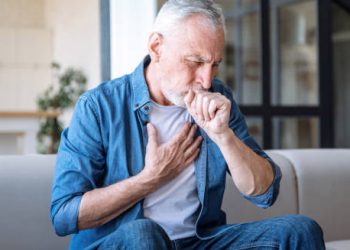 Older man coughing and clutching chest while sitting, showing symptoms of heart disease