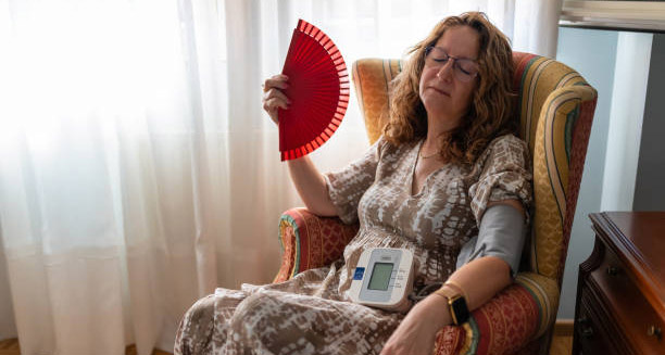 Woman fanning herself while monitoring blood pressure, experiencing discomfort from heart rhythm symptoms