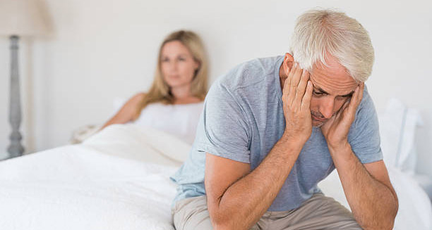 Older man sitting on bed with head in hands, showing symptoms of impotence