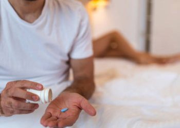 Man taking blue pill for impotence treatment while partner lies in bed