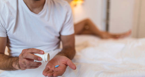 Man taking blue pill for impotence treatment while partner lies in bed